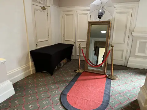 A full-length framed mirror stands behind a red and black floor runner, with red ropes and an umbrella light.