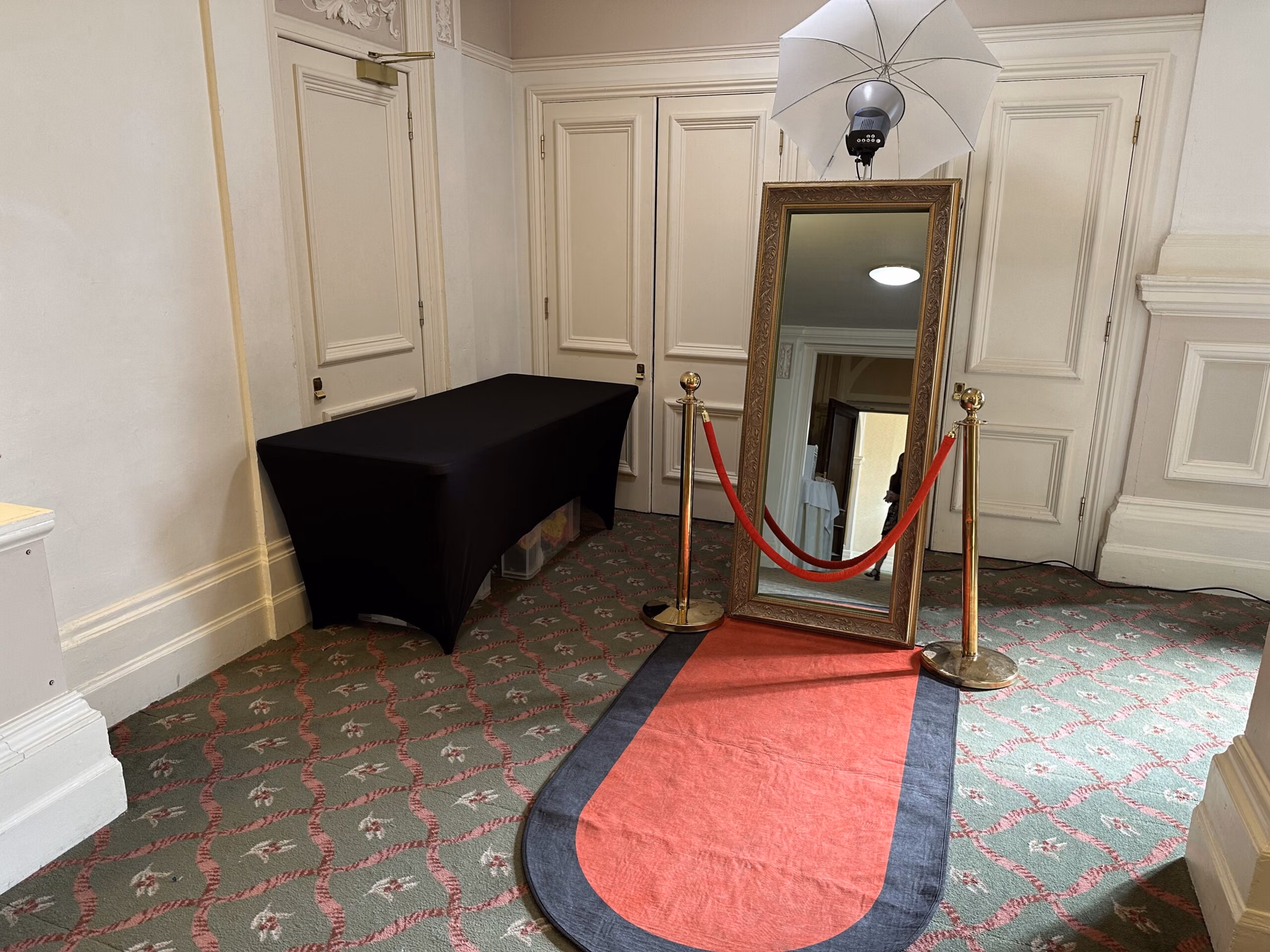 A full-length framed mirror stands behind a red and black floor runner, with red ropes and an umbrella light.