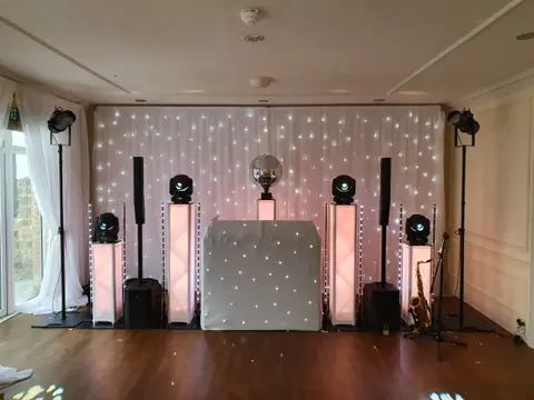 A lit DJ booth with speakers, uplights and a starry curtain backdrop in a room