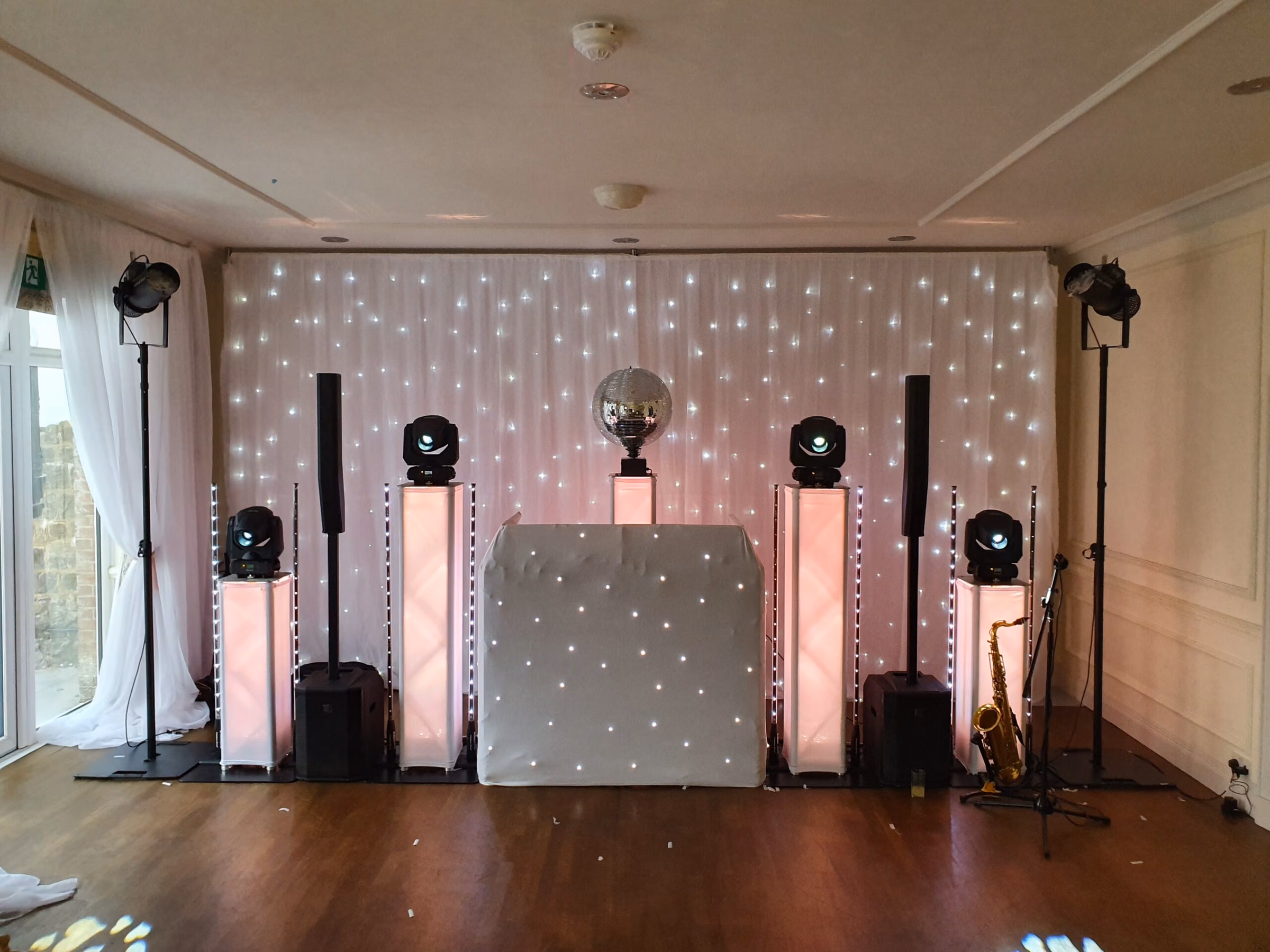 A lit DJ booth with speakers, uplights and a starry curtain backdrop in a room