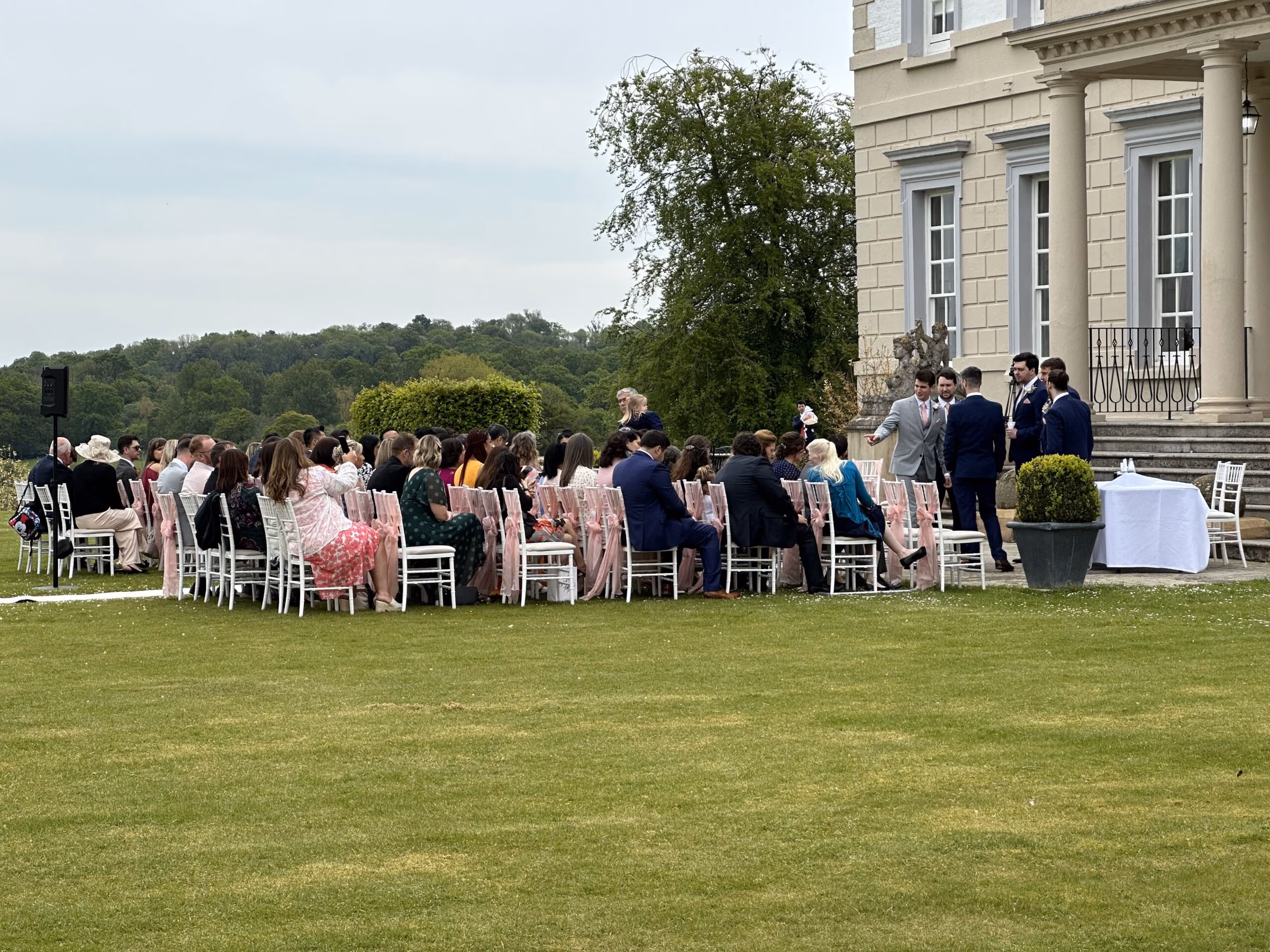 Guests seated for an outdoor wedding ceremony beside a country house, with a speaker at the side.
