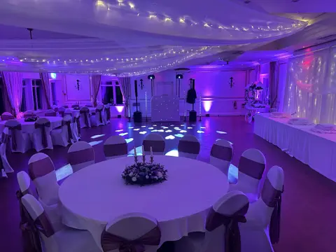 Purple-lit banquet hall with round tables, white chairs, draped ceiling fabric and floral centrepieces.