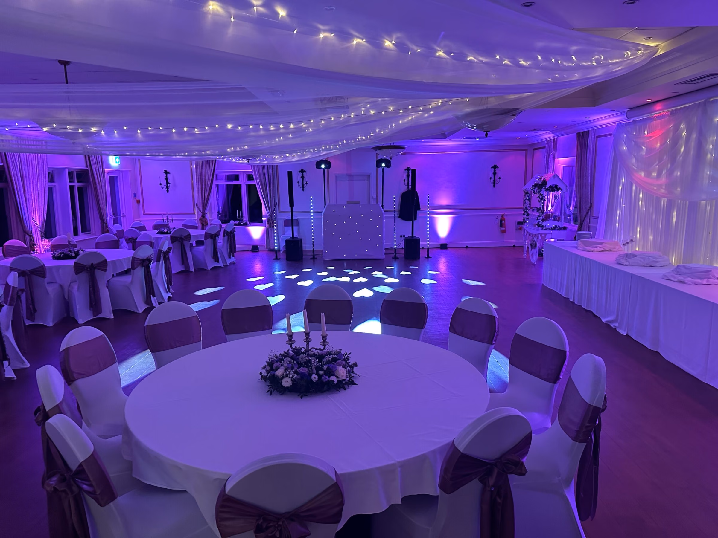 Purple-lit banquet hall with round tables, white chairs, draped ceiling fabric and floral centrepieces.