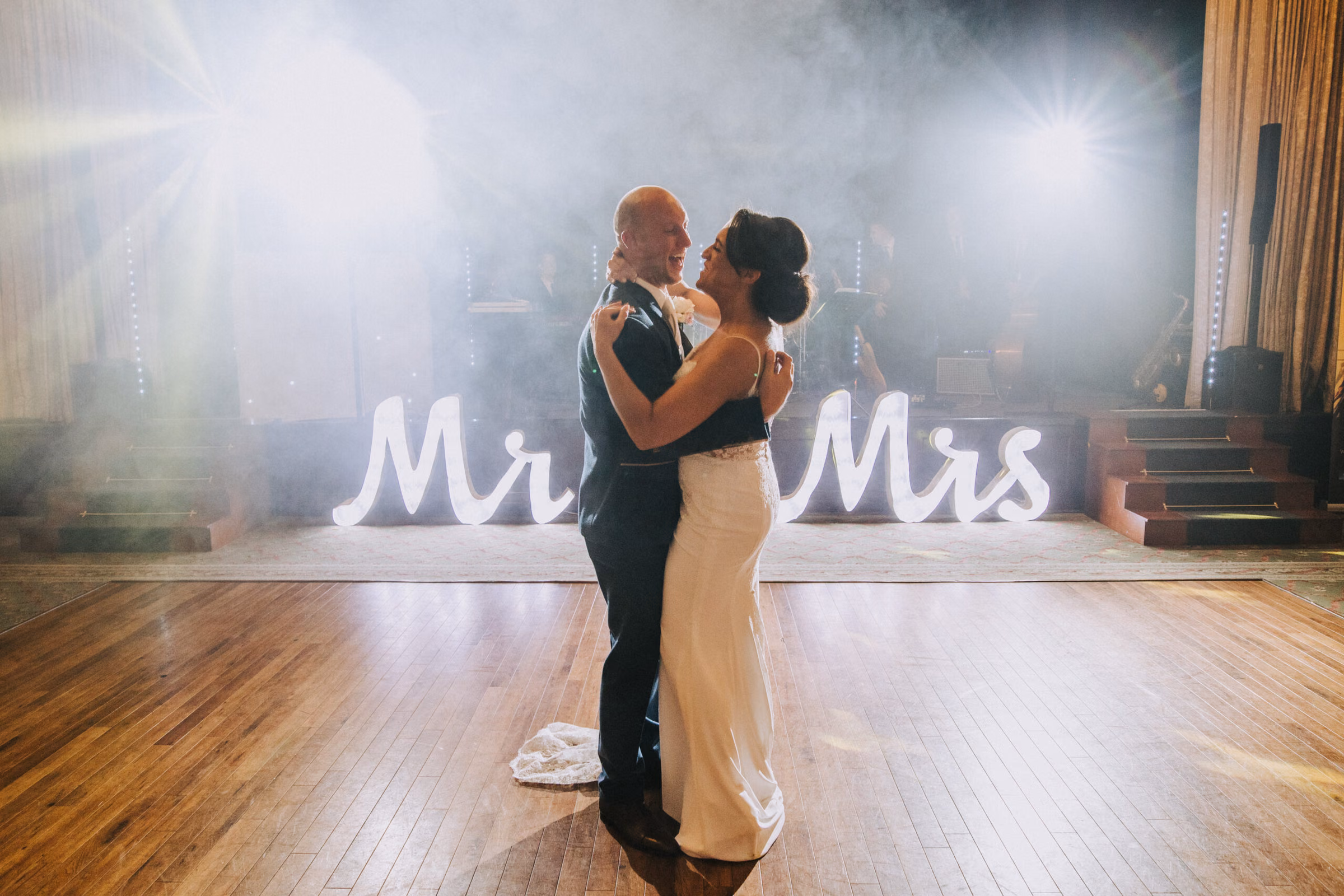 A wedding couple dancing on a wooden floor with illuminated Mr & Mrs letters behind them.